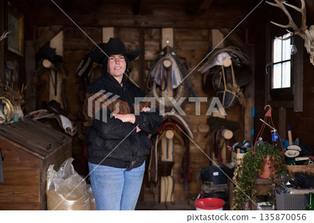 Cowgirl in a hat stands with arms crossed inside a rustic tack room filled with saddles 135870056