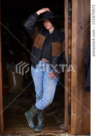 Cowgirl in doorway at rustic ranch wearing hat, jeans, and boots 135870072