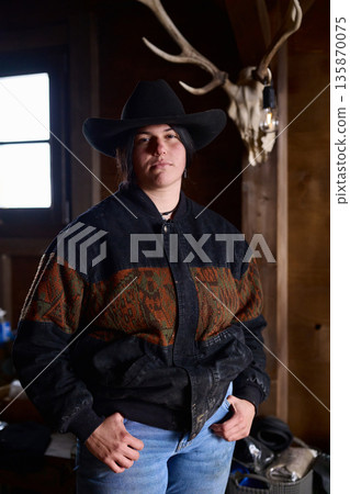 Cowgirl in a wide brimmed hat stands confidently in a rustic ranch interior Cowgirl in a wide brimmed hat stands confidently in a rustic ranch interior 135870075
