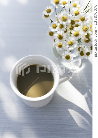 陽光透過樹木灑在白桌上,咖啡和母菊擺放在桌上(垂直,留白) 陽光透過樹木灑在白桌上,咖啡和母菊擺放在桌上(垂直,留白) 135870076