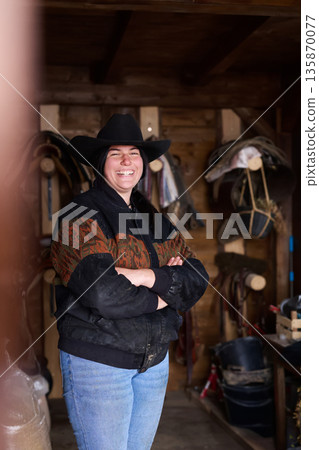 Happy cowgirl in a black hat stands in a wooden barn with tools, smiling confidently Happy cowgirl in a black hat stands in a wooden barn with tools, smiling confidently 135870077