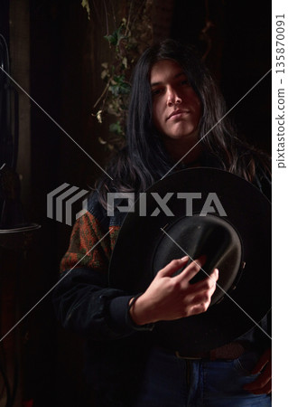 Moody portrait of a young woman holding a wide-brim hat in a dark rustic setting Moody portrait of a young woman holding a wide-brim hat in a dark rustic setting 135870091