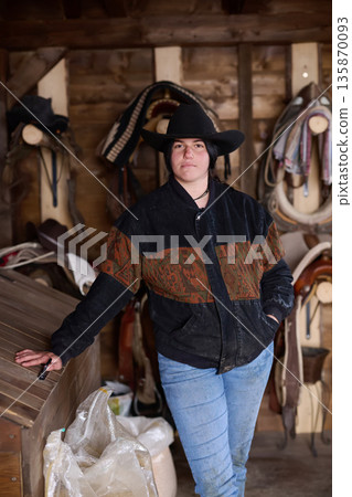 Cowgirl in a black hat and patterned jacket stands in a horse tack room at a ranch 135870093