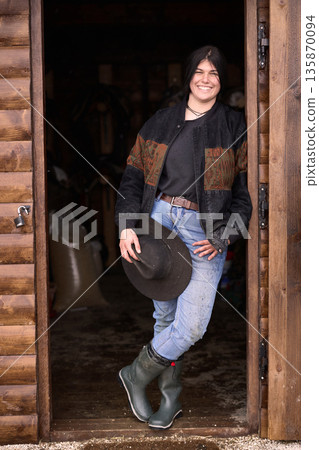 Smiling cowgirl stands in rustic barn doorway wearing jacket, jeans, and hat Smiling cowgirl stands in rustic barn doorway wearing jacket, jeans, and hat 135870094