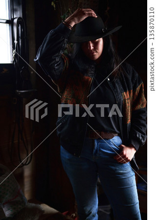 Cowgirl in black hat and denim jacket poses in a dimly lit room with casual confidence Cowgirl in black hat and denim jacket poses in a dimly lit room with casual confidence 135870110