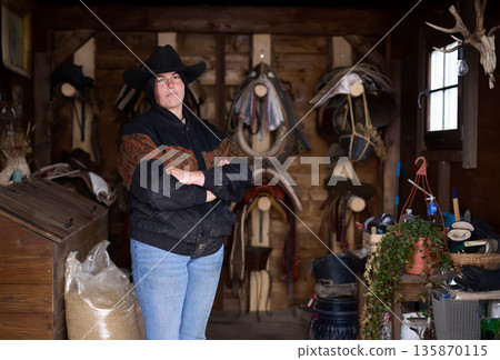 Cowgirl in a ranch workshop standing with arms crossed among saddles and gear 135870115