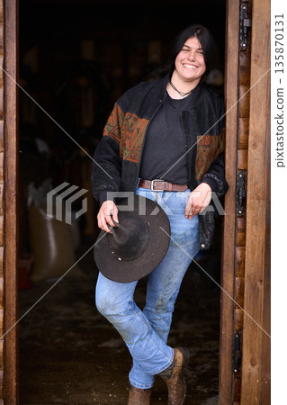 Smiling cowgirl stands in a wooden doorway with a hat, jeans, and boots at the ranch Smiling cowgirl stands in a wooden doorway with a hat, jeans, and boots at the ranch 135870131
