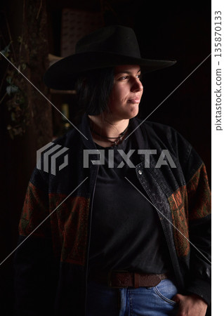 Cowgirl in a dark hat and plaid jacket gazes off to the side on a ranch setting 135870133