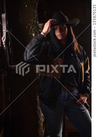 Cowgirl in a rustic ranch setting adjusting her hat in denim jacket and jeans Cowgirl in a rustic ranch setting adjusting her hat in denim jacket and jeans 135870135