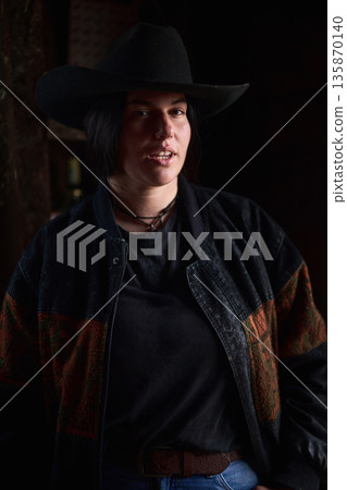 Cowgirl in a black hat and denim jacket on a dimly lit ranch, confident and stylish. 135870140
