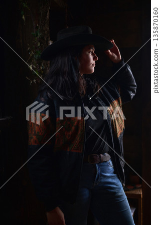 Cowgirl in a wide brim hat and patterned jacket stands by a doorway in moody, rustic light Cowgirl in a wide brim hat and patterned jacket stands by a doorway in moody, rustic light 135870160