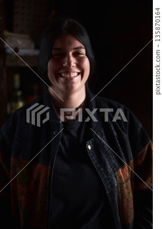 Smiling woman in a dark jacket stands against a dimly lit background, conveying warmth and casual style Smiling woman in a dark jacket stands against a dimly lit background, conveying warmth and casual style 135870164
