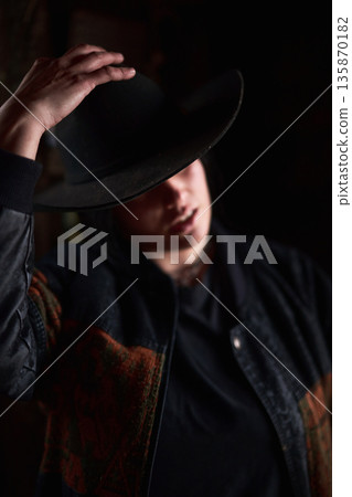 Person tipping a wide-brim hat in a moody western portrait wearing a denim jacket 135870182