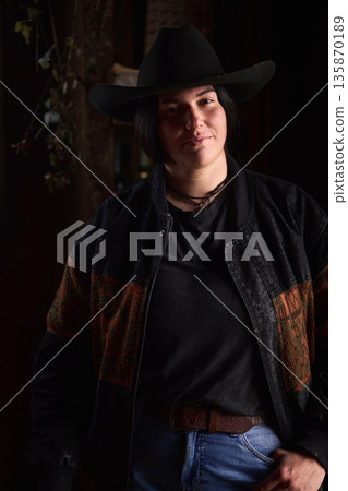 Cowgirl in black hat and denim, posing in a dark rustic setting at the ranch Cowgirl in black hat and denim, posing in a dark rustic setting at the ranch 135870189