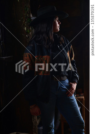 Cowgirl in wide hat and denim, standing in a moody ranch setting with a confident pose Cowgirl in wide hat and denim, standing in a moody ranch setting with a confident pose 135870191