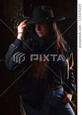 Cowgirl in Wide Hat and Dark Jacket Stands by Rustic Wood Wall, Moody Western Portrait Cowgirl in Wide Hat and Dark Jacket Stands by Rustic Wood Wall, Moody Western Portrait 135870205