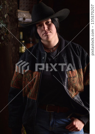 Cowgirl in a black cowboy hat and denim jacket poses confidently in a rustic, dimly lit setting Cowgirl in a black cowboy hat and denim jacket poses confidently in a rustic, dimly lit setting 135870207