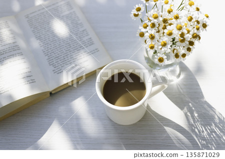 A white table with sunlight filtering through the trees, reading, coffee, and matricaria (horizontal, white space) A white table with sunlight filtering through the trees, reading, coffee, and matricaria (horizontal, white space) 135871029