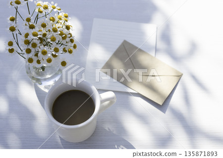 A white table in the sunlight filtering through the trees: coffee, matricaria, and a letter (envelope, letter paper, and blank space) A white table in the sunlight filtering through the trees: coffee, matricaria, and a letter (envelope, letter paper, and blank space) 135871893
