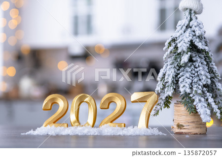 Golden number 2027 on table in background of cozy white kitchen with Christmas tree and Christmas decor with garland lights. Housing, interior, renovation, mortgage, rent in New Year 135872057