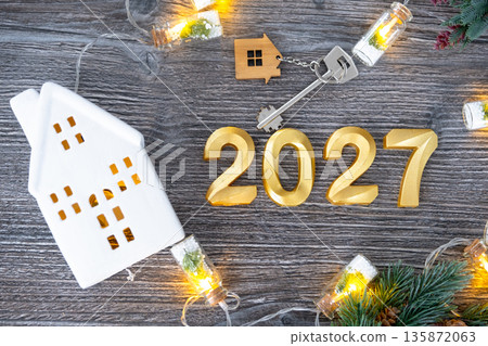 New Year 2027 golden numbers and House key with tiny form of home on festive grey wooden background flatlay, lights of garlands, greeting card. Housing, real estate, relocation, mortgage, insurance 135872063