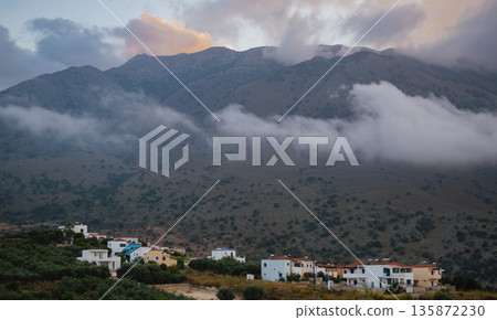 Traditional houses in a village at the foot of foggy, cloud-covered mountains in Crete Traditional houses in a village at the foot of foggy, cloud-covered mountains in Crete 135872230