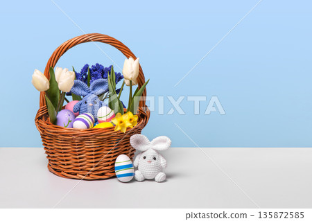 Multicolored Easter eggs in a basket with spring flowers: blue muscari, yellow daffodils and white tulips and white Easter knitted bunny with blue egg on gray table. Easter concept Multicolored Easter eggs in a basket with spring flowers: blue muscari, yellow daffodils and white tulips and white Easter knitted bunny with blue egg on gray table. Easter concept 135872585