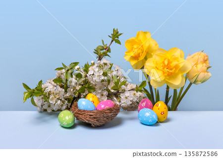 Multicolored Easter eggs in bird nest with spring flowers: blooming spring cherry branches and yellow daffodils on gray table. Easter concept Multicolored Easter eggs in bird nest with spring flowers: blooming spring cherry branches and yellow daffodils on gray table. Easter concept 135872586
