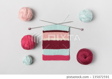 Brown beige vinous and turquoise piece of striped knitting with knitting needles and balls of thread on gray background. Homemade needlework and knitting of warm, clothes and toys. Concept creativity 135872591