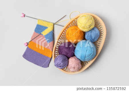 Set for the hobby of knitting warm clothes or toys. Yellow, green, blue, orange and lilac colors knitting balls in basket with knitting needles and knitted scarf on gray background. Concept needlework Set for the hobby of knitting warm clothes or toys. Yellow, green, blue, orange and lilac colors knitting balls in basket with knitting needles and knitted scarf on gray background. Concept needlework 135872592