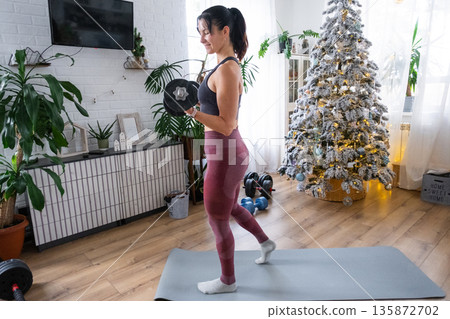 Woman in sports uniform is engaged in fitness and strength training with dumbbells at home in Christmas interior with Christmas tree. Preparation and Recovery of athletic form after New Year Woman in sports uniform is engaged in fitness and strength training with dumbbells at home in Christmas interior with Christmas tree. Preparation and Recovery of athletic form after New Year 135872702