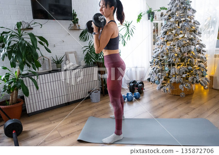 Woman in sports uniform is engaged in fitness and strength training with dumbbells at home in Christmas interior with Christmas tree. Preparation and Recovery of athletic form after New Year Woman in sports uniform is engaged in fitness and strength training with dumbbells at home in Christmas interior with Christmas tree. Preparation and Recovery of athletic form after New Year 135872706