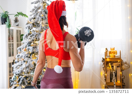 Woman in sports uniform and Santa hat is engaged in fitness and strength training with dumbbells at home in Christmas interior with Christmas tree. Recovery of athletic form after New Year 135872711
