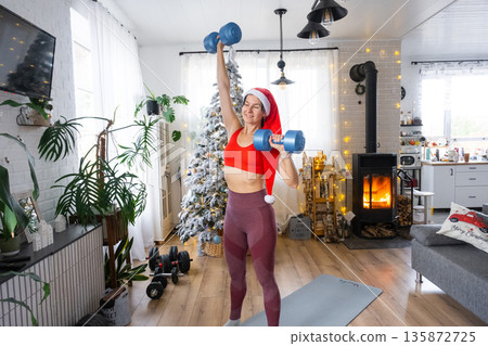 Woman in sports uniform and Santa hat is engaged in fitness and strength training with dumbbells at home in Christmas interior with Christmas tree. Recovery of athletic form after New Year Woman in sports uniform and Santa hat is engaged in fitness and strength training with dumbbells at home in Christmas interior with Christmas tree. Recovery of athletic form after New Year 135872725