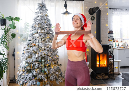 Woman in sports uniform and Santa hat is engaged in fitness and strength training with fitness elastic bands at home in Christmas interior with Christmas tree. Recovery of athletic form after New Year Woman in sports uniform and Santa hat is engaged in fitness and strength training with fitness elastic bands at home in Christmas interior with Christmas tree. Recovery of athletic form after New Year 135872727