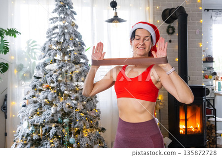 Woman in sports uniform and Santa hat is engaged in fitness and strength training with fitness elastic bands at home in Christmas interior with Christmas tree. Recovery of athletic form after New Year Woman in sports uniform and Santa hat is engaged in fitness and strength training with fitness elastic bands at home in Christmas interior with Christmas tree. Recovery of athletic form after New Year 135872728