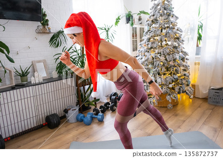 Woman in sports uniform and Santa hat is engaged in fitness and strength training with fitness elastic bands at home in Christmas interior with Christmas tree. Recovery of athletic form after New Year 135872737