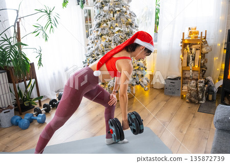 Woman in sports uniform and Santa hat is engaged in fitness and strength training with dumbbells at home in Christmas interior with Christmas tree. Recovery of athletic form after New Year Woman in sports uniform and Santa hat is engaged in fitness and strength training with dumbbells at home in Christmas interior with Christmas tree. Recovery of athletic form after New Year 135872739