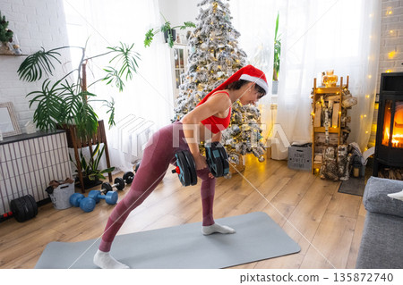 Woman in sports uniform and Santa hat is engaged in fitness and strength training with dumbbells at home in Christmas interior with Christmas tree. Recovery of athletic form after New Year Woman in sports uniform and Santa hat is engaged in fitness and strength training with dumbbells at home in Christmas interior with Christmas tree. Recovery of athletic form after New Year 135872740