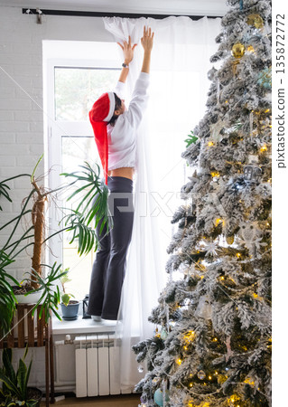 A woman hang up white curtain on the window, preparation for the Christmas and New Year, fairy lights in the interior of a house with a Christmas tree A woman hang up white curtain on the window, preparation for the Christmas and New Year, fairy lights in the interior of a house with a Christmas tree 135872772