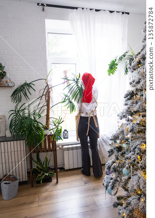 Woman in a Santa hat and apron cleans window near Christmas tree, preparing for the New Year, decorating and tidying up the house 135872784