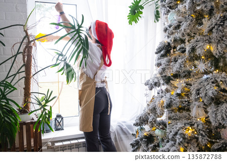 Woman in a Santa hat and apron cleans window near Christmas tree, preparing for the New Year, decorating and tidying up the house 135872788