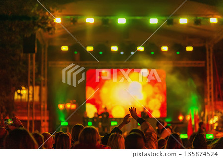 Crowd with hands enjoying live music concert with vibrant stage lights and colorful LED screen in background, music festival. Concept of concert, music, entertainment, nightlife celebration 135874524
