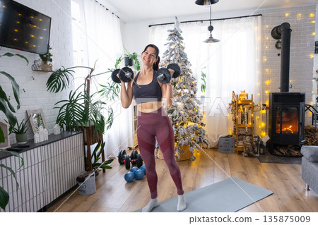 Woman in sports uniform is engaged in fitness and strength training with dumbbells at home in Christmas interior with Christmas tree. Preparation and Recovery of athletic form after New Year 135875009