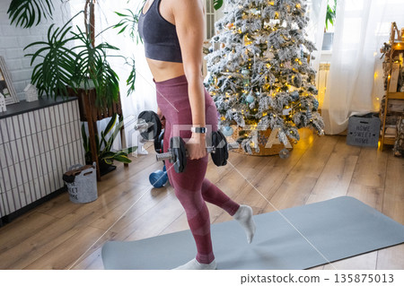 Woman in sports uniform is engaged in fitness and strength training with dumbbells at home in Christmas interior with Christmas tree. Preparation and Recovery of athletic form after New Year Woman in sports uniform is engaged in fitness and strength training with dumbbells at home in Christmas interior with Christmas tree. Preparation and Recovery of athletic form after New Year 135875013