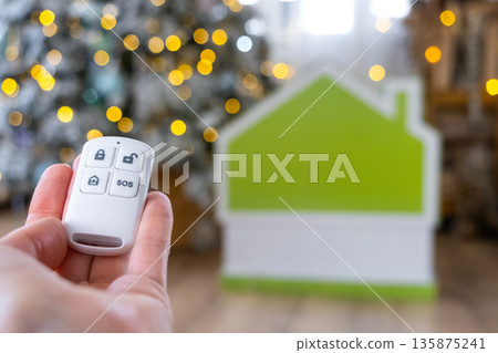 Security System Keychain in hand on background fairy lights Christmas tree and tiny house gift for New Year, Christmas. Home security, alarm system, SOS signal, protection from thieves Security System Keychain in hand on background fairy lights Christmas tree and tiny house gift for New Year, Christmas. Home security, alarm system, SOS signal, protection from thieves 135875241