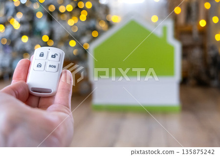 Security System Keychain in hand on background fairy lights Christmas tree and tiny house gift for New Year, Christmas. Home security, alarm system, SOS signal, protection from thieves Security System Keychain in hand on background fairy lights Christmas tree and tiny house gift for New Year, Christmas. Home security, alarm system, SOS signal, protection from thieves 135875242