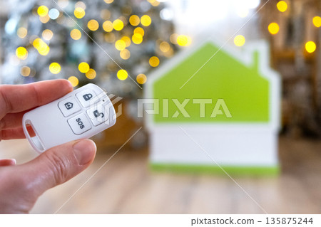 Security System Keychain in hand on background fairy lights Christmas tree and tiny house gift for New Year, Christmas. Home security, alarm system, SOS signal, protection from thieves Security System Keychain in hand on background fairy lights Christmas tree and tiny house gift for New Year, Christmas. Home security, alarm system, SOS signal, protection from thieves 135875244