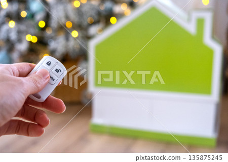 Security System Keychain in hand on background fairy lights Christmas tree and tiny house gift for New Year, Christmas. Home security, alarm system, SOS signal, protection from thieves 135875245