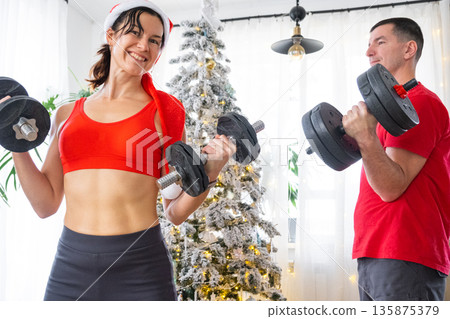 Woman and man in sports uniform and Santa hat is engaged in fitness and strength training with dumbbells at home in Christmas interior with Christmas tree. Recovery of athletic form after the New Year Woman and man in sports uniform and Santa hat is engaged in fitness and strength training with dumbbells at home in Christmas interior with Christmas tree. Recovery of athletic form after the New Year 135875379